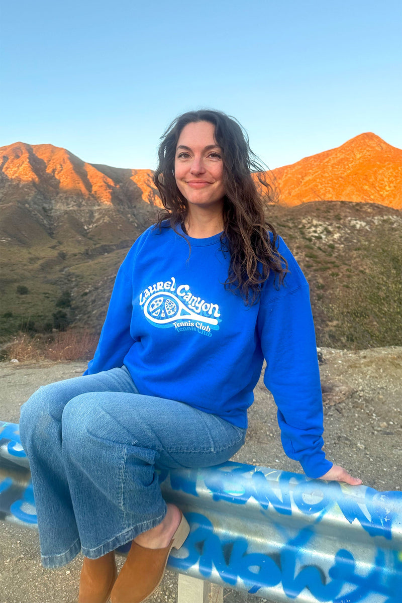 Person wearing a blue sweatshirt with a logo, sitting on a guardrail with mountains in the background.
